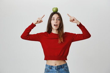 Attractive young female in red crop top holding apple on head, pointing with index fingers expressing surprise, isolated over white background. She lost so girl need to stand like this till fruit fall