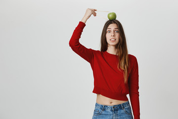 Portrait of young funny fashionable girl in trendy cropped top trying to grab apple placed on her head with chopsticks, isolated over white background. Friends made up such punishment for being late