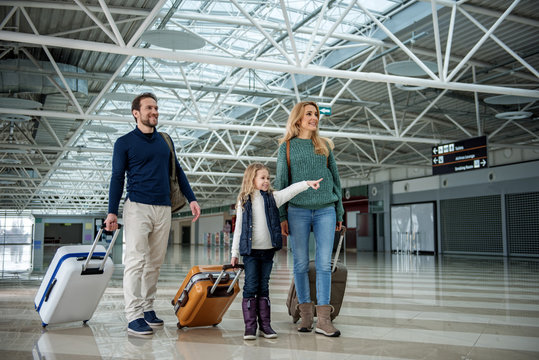 Cheerful parents and their daughter standing with luggage at the airport. Kid is pointing with finger