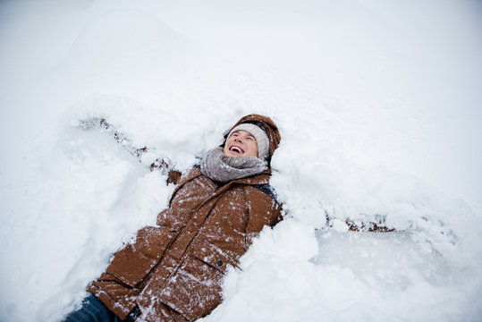 Top View Of Excited Young Man Lying On Snow While Male Angel Wings By Arms. He Is Looking Up And Laughing. Enjoying Winter Concept