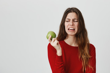 Portrait of young european teenager expressing disgust holding green apple, isolated over white background. People say fruit keep doctors away, but girl still don't like it.