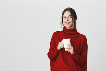 Portrait of young attractive european student with shiny smile, wearing stylish red sweater holding a cup while talking to barista at local cafe. Girl will write about it in her blog