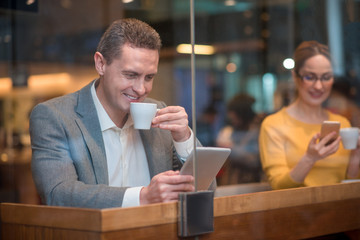 Portrait of cheerful male watching at electronic tablet. Happy girl typing in mobile. They tasting mugs of tea while situating at desk. Relax concept