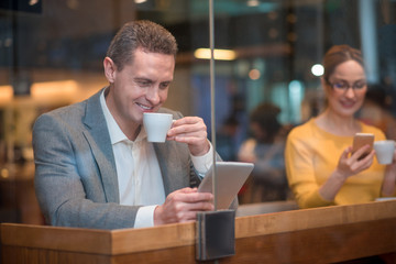 Portrait of cheerful male watching at electronic tablet. Happy girl typing in mobile. They tasting mugs of tea while situating at desk. Relax concept