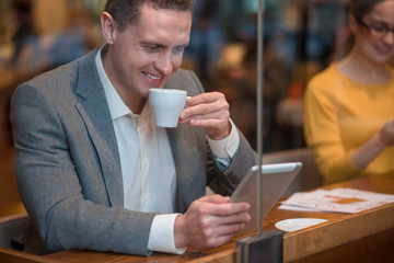 Outgoing man drinking cup of appetizing coffee while looking at digital device. He sitting near smiling female at table in cafe. Rest concept
