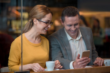 Cheerful female and smiling male looking at mobile while tasting mug of beverage. They sitting at table in cafe. Relax concept