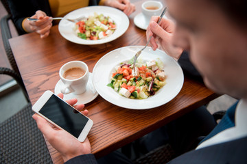 Close up man hand typing in mobile while tasting appetizing dishes. Colleague locating opposite him. Business break concept