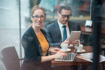 Portrait of cheerful businesswoman typing in laptop while sitting at desk. Calm colleague looking at electronic tablet. Occupation concept