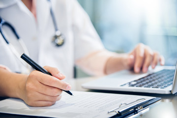 female doctor is using laptop and writing something on clipboard.