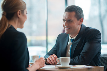 Outgoing man telling with cheerful female comrade while tasting mug of beverage. Business meeting concept