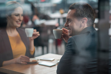 Side view outgoing man tasting cups of liquid while locating at desk in cafeteria. He telling with happy female. Coffee break concept