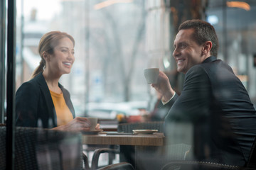 Laughing businesswoman telling with beaming male affiliate while situating at tale in cafe. They drinking mugs of beverage. Leisure concept