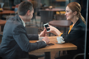 Happy female pointing on screen of mobile while sitting at desk in cafe. Male partner looking at it. Leisure and job concept