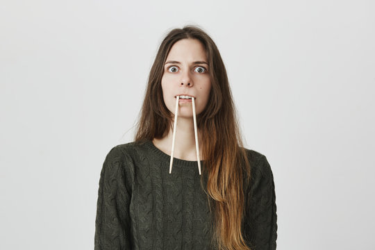 Portrait of funny childish adult female holding chopsticks in mouth imitating mammoth or beaver over white background. Girl plays guess game with friends trying to help them name animal she shows.