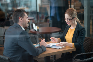 Portrait of outgoing young woman writing in appointment book while sitting at desk. She speaking with smiling comrade. Communication and job concept