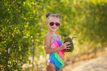 Little girl in a swimsuit and sunglasses on the beach with a cocktail in pineapple