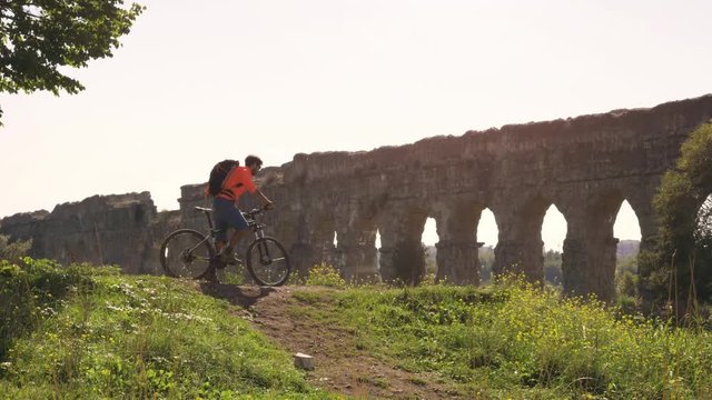 Cyclist riding bike in front of ancient roman aqueduct in a park. Young attractive athletic man with orange sportswear and backpack in parco degli acquedotti in Rome
