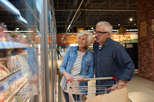 Portrait of modern senior couple grocery shopping in supermarket, smiling happily while choosing frozen foods standing by freezers , copy space
