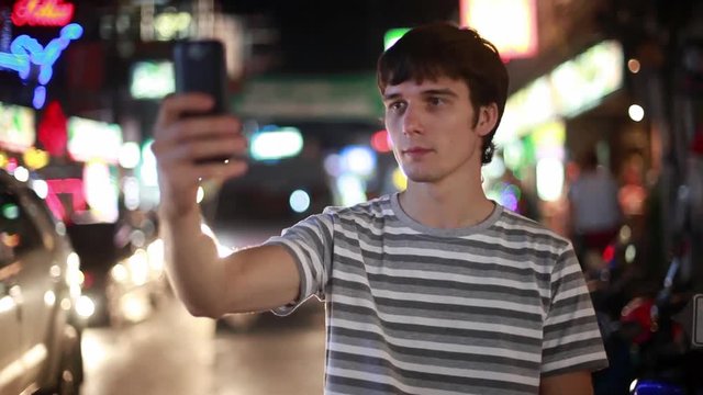 Tourist man with mobile phone takes picture in the night street with bokeh lights. 1920x1080