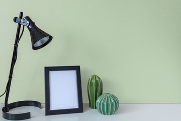 Aa white desk with a black lamp, a frame with space for text and two green cactuses on the background of a light green wall