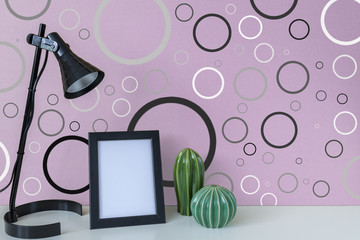 A white desk with a black lamp, a frame with space for text and two green cactuses on the background of a light pink wall in circles