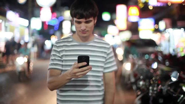 Portrait of handsome man walks, smiles, uses his smartphone and looks at camera. Blurred busy city lights in the street. Modern technology. 1920x1080