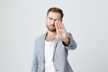 Serious confident angry european man with beard wearing stylish clothes posing against gray studio wall, keeping hands in stop gesture, as if saying: Stay away from me. Body language.