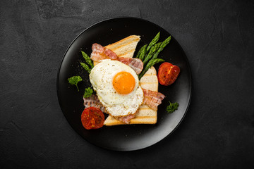 Overhead shot of breakfast or lunch with fried egg, bread toast, green asparagus, tomatoes and bacon on black plate with copy space.
