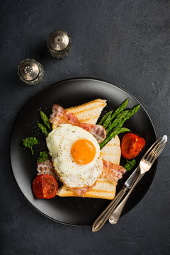 Breakfast or lunch with Fried egg, bread toast, green asparagus, tomatoes and bacon on black plate. Top view. Copy space.