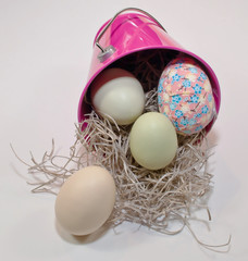 Easter eggs shown spilling out of a pink bucket along with straw on a white background