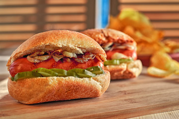 Hot dog with fried onions and marinated cucumbers. Potato chips. Classic fast food on wooden table. Warm glow.