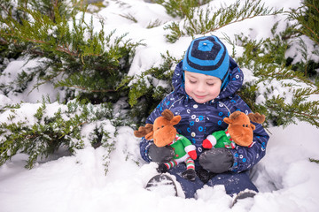 adorable preschooler in winter wear sit amoung snow and play with toy reindeer.