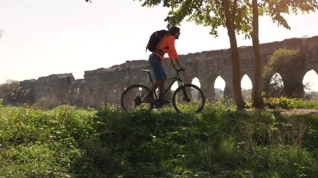 Cyclist riding bike in front of ancient roman aqueduct in a park. Young attractive athletic man with orange sportswear and backpack in parco degli acquedotti in Rome
