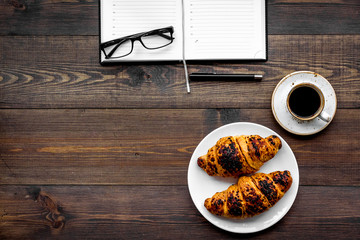 Work at breakfast. Coffee and croissants near notebook and glasses. Dark wooden background top view copy space