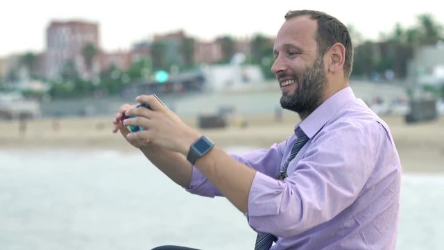 Happy businessman taking selfie photo with cellphone sitting on rock near sea 

