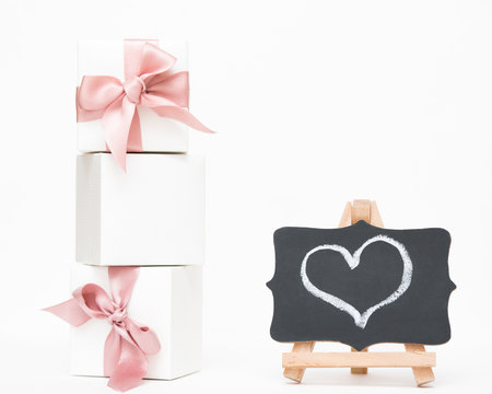 White gift boxes of the famous jewelry brand with bracelets and charms with a pink ribbon. Mock up. Empty place for an inscription. Postcard for the holiday. Heart drawn chalk on a blackboard.