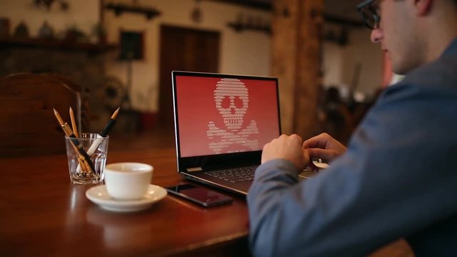 Man turns on a laptop waits for loading computer and finds out it is infected by a ransomware spyware virus that is asking for money to retrieve encrypted data. Scary red skull crossbones on screen.