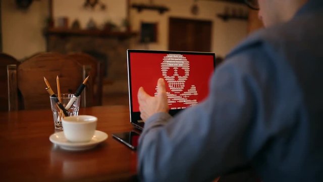 Man with a laptop being infected by a ransomware spyware virus that is asking for money to retrieve the encrypted files. Scary red shimmering skull and crossbones on the screen.