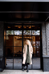 Portrait of beautiful young stylish blonde woman wearing beige coat and walking through the city streets. Trendy casual outfit. Street fashion.