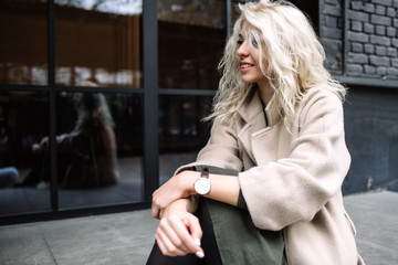 Fototapeta premium Portrait of beautiful young stylish blonde woman wearing beige coat and walking through the city streets. Trendy casual outfit. Street fashion.