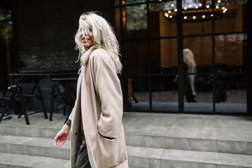 Naklejka premium Portrait of beautiful young stylish blonde woman wearing beige coat and walking through the city streets. Trendy casual outfit. Street fashion.