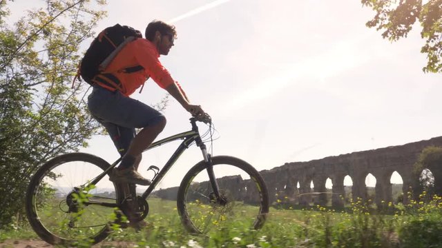 Cyclist riding bike in front of ancient roman aqueduct in a park. Young attractive athletic man with orange sportswear and backpack in parco degli acquedotti in Rome
