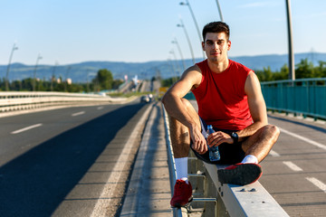 Young male runner drinking water and resting after morning workout on bridge.