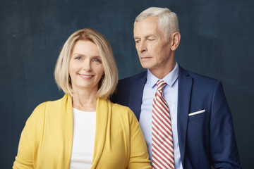 Studio shot of senior couple standing at dark background. Smiling mature woman looking at camera and elderly man wearing suit while standing next to her. 