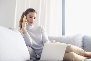 Managing her business from home. An attractive middle aged woman using her cell phone and making call while sitting on sofa with laptop and working from home office.