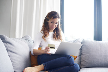 Banking from home is comfy. A woman sitting on sofa and looking thoughtful while shopping online with her credit card.