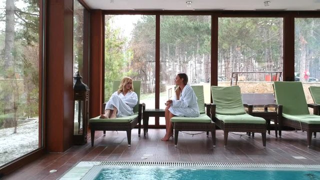 Young women relaxing together in beautiful spa laying on wooden chair near the pool
