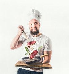 Young cheerful cook man with beard holding a frying pan with steaks and knife flying in the air on light background. Meat food and cooking concept