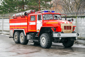 The fire truck in full equipment rushes to a challenge along the asphalt road next to the concrete fence.