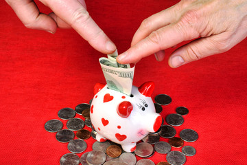Romantic ceramic piggy bank over a lot of coins with a red background. Human fingers pushed the...